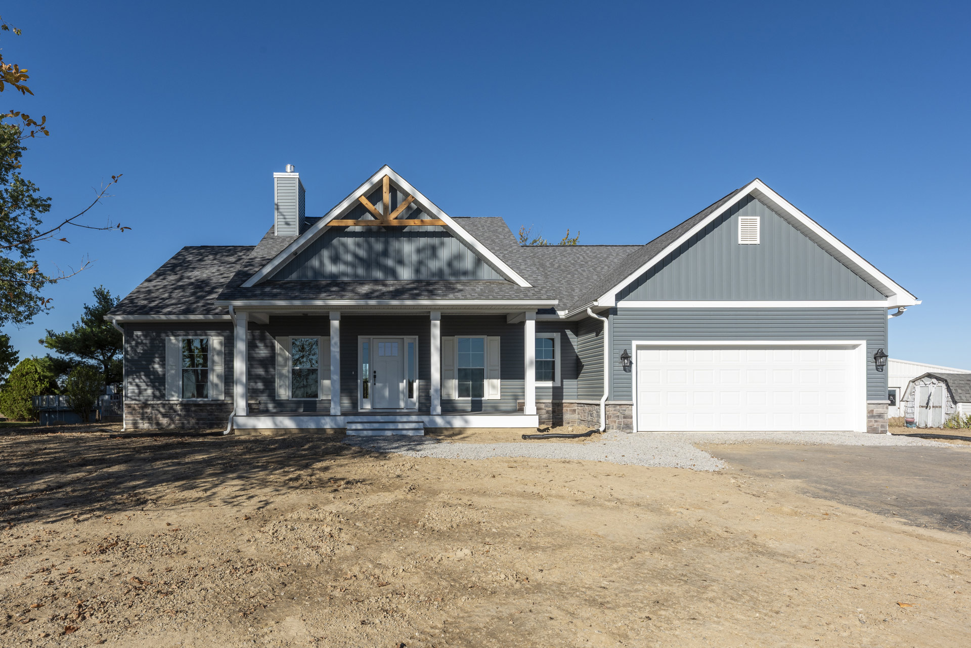 Two-story home with white siding, gray shingle roof, white garage door, paved driveway, multi-pane windows reflecting trees, and dirt area with black pipe in front.