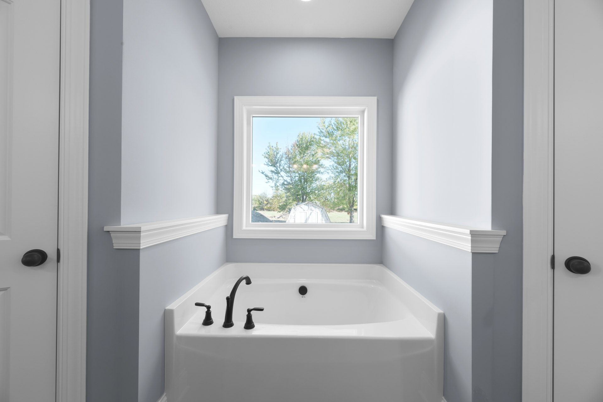 Freestanding white bathtub beneath large window overlooking trees and shed, chrome faucet mounted on tiled wall