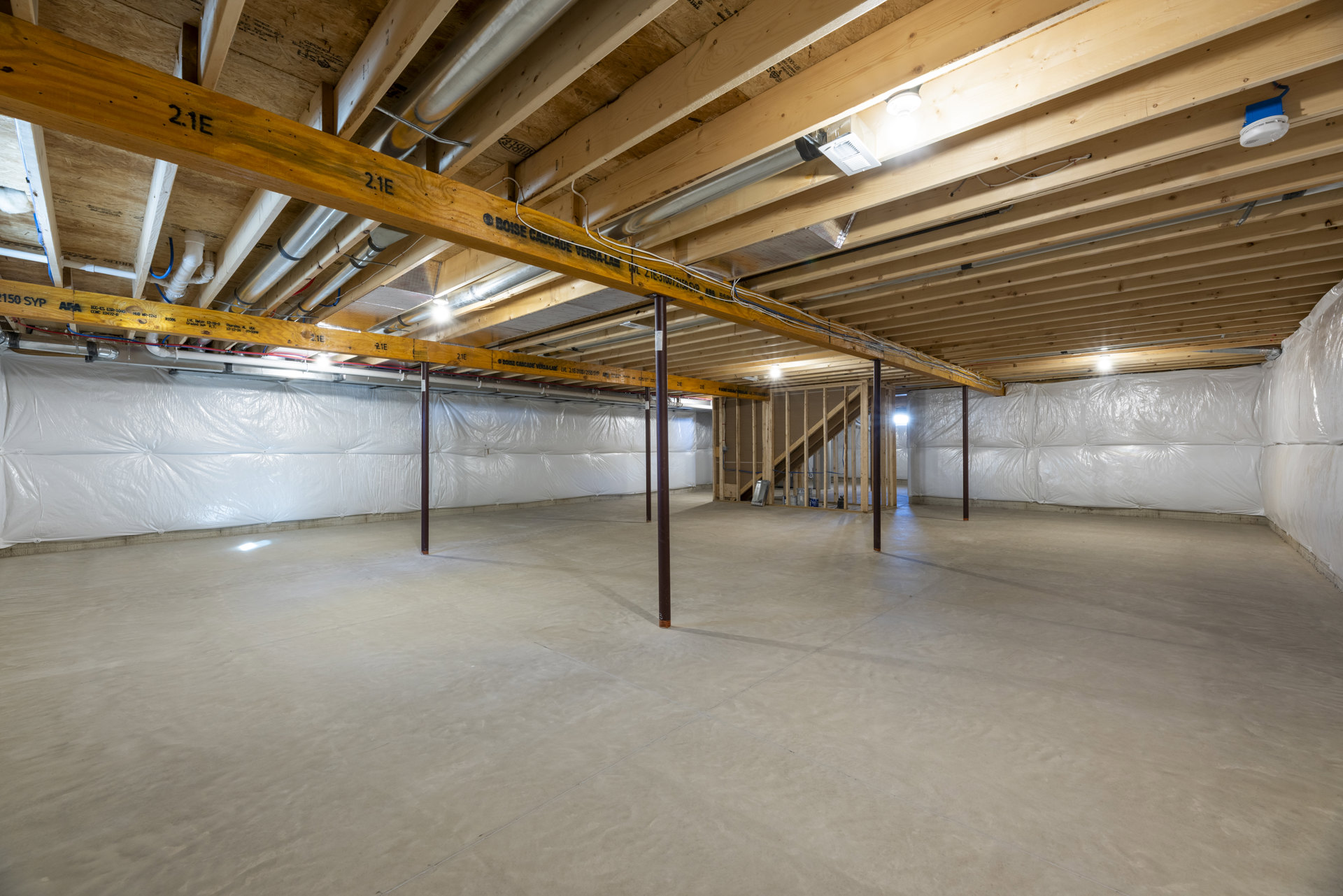 Exposed wooden beams and metal pipes across a white wall, concrete floor, and structural pole in a basement room