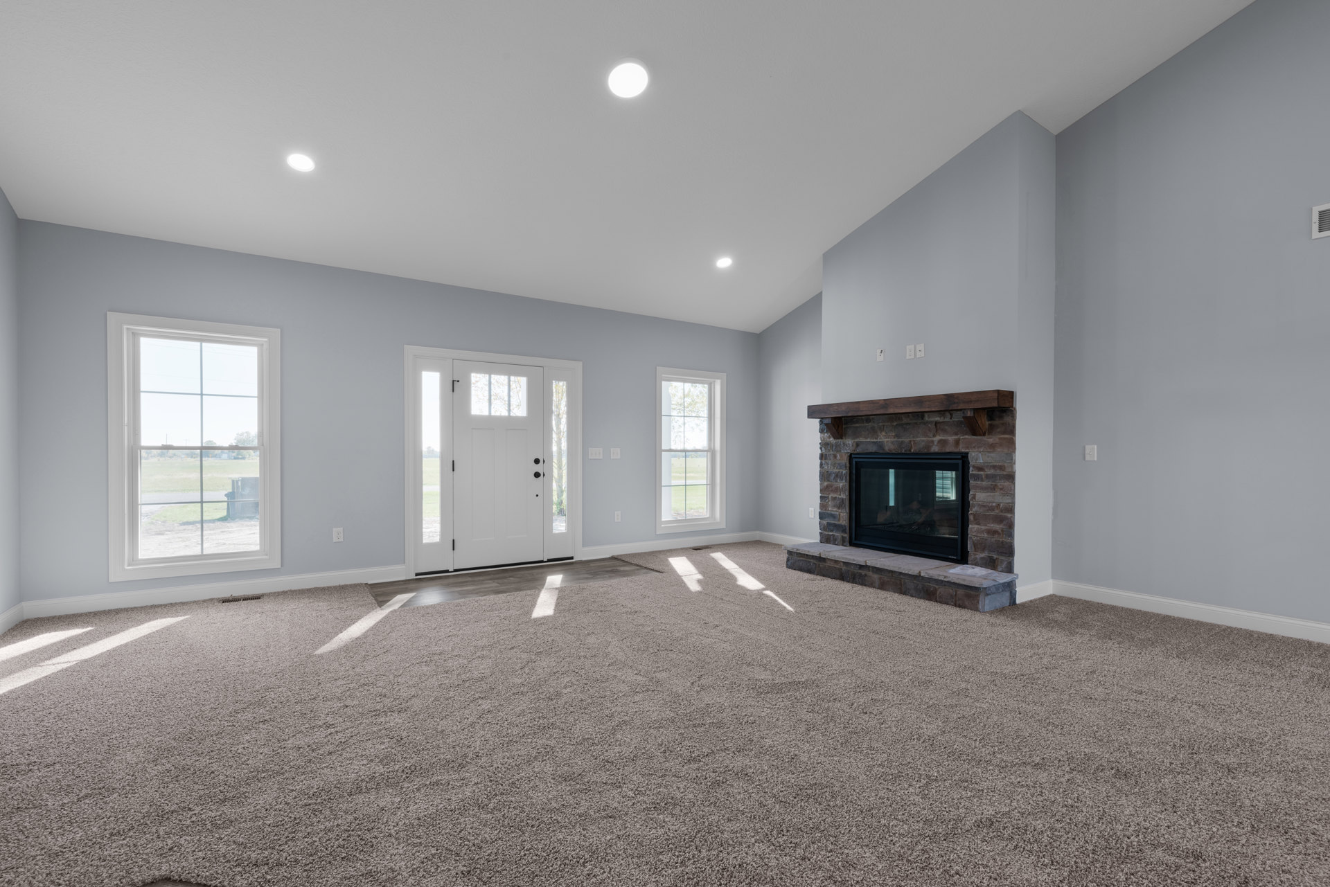 Living room with carpeted floor, central fireplace featuring a glass door and wood mantel, large window overlooking a field, white door with glass panels, plaster walls and