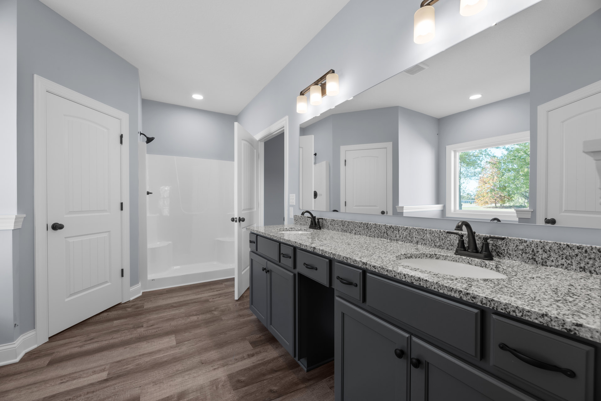 Bathroom featuring marble countertop, white cabinetry, undermount sink, glass-enclosed shower, white door with black hardware, window overlooking trees, and recessed ceiling light