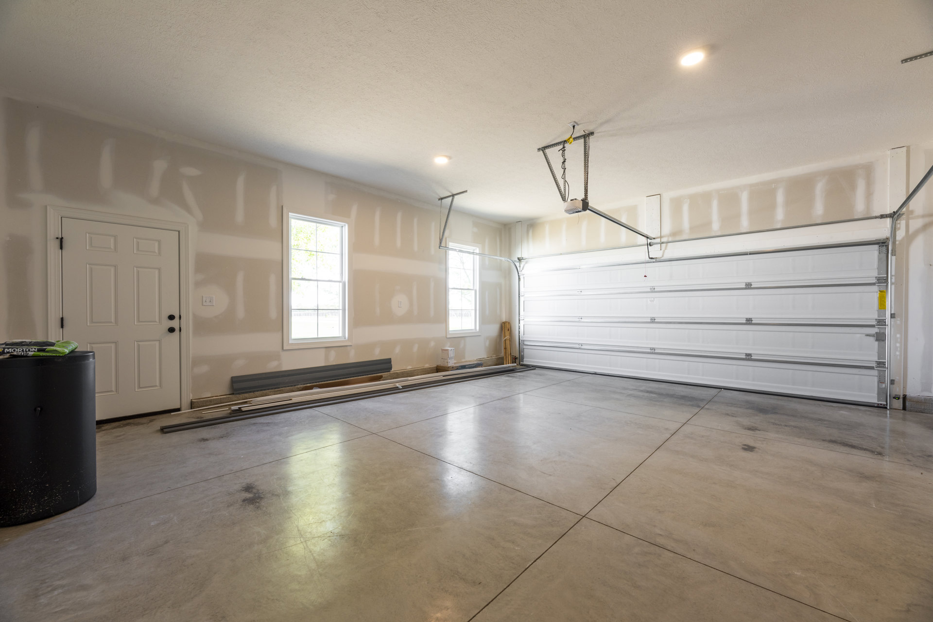 Garage interior featuring white plaster walls, white paneled garage door, ceiling-mounted light fixture, light switch on the wall, gray flooring, window with white frame, and black