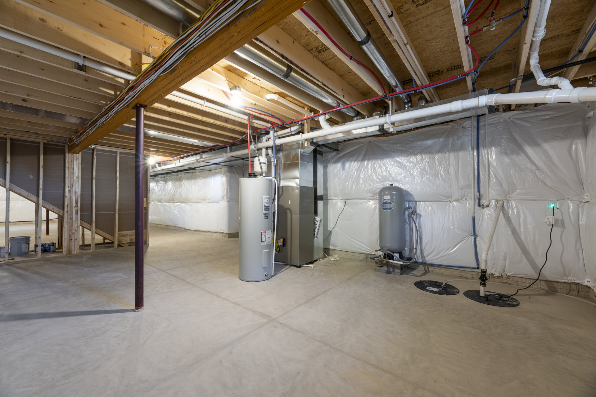 Basement utility room with exposed steel beams, insulated ceiling, large white cylindrical tank labeled on the side, dense network of pipes and wires overhead, composite flooring