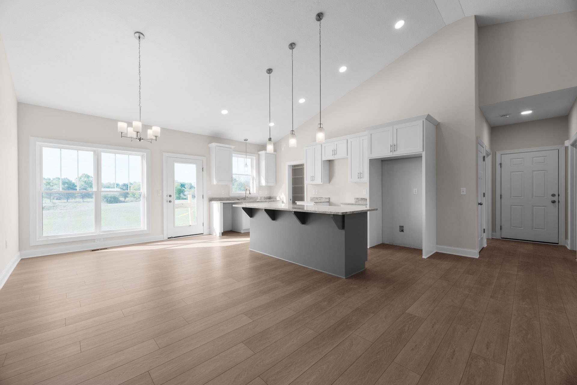 Kitchen with wood flooring, large window overlooking grassy field, white cabinetry, grey walls, and white door with silver handle