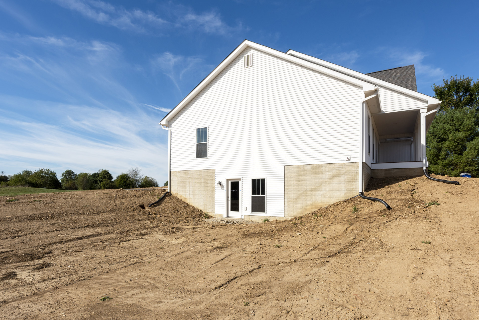 White house with white roof and white-framed windows, set beside a large dirt hill under a blue sky with scattered clouds.