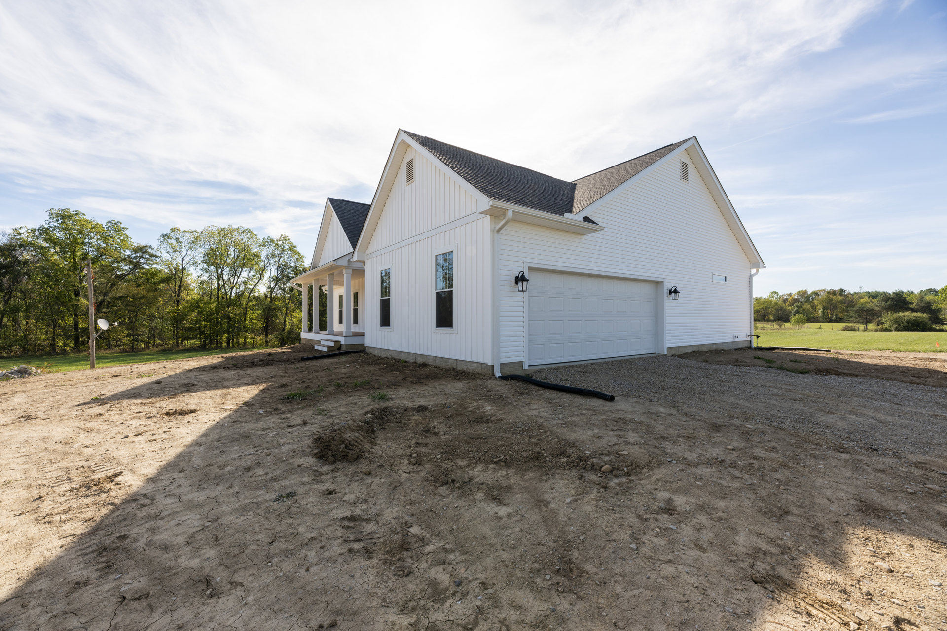 White house with black roof and attached garage, dirt patch and rocks in front yard, black pipe on ground, satellite dish on pole near wooded area.