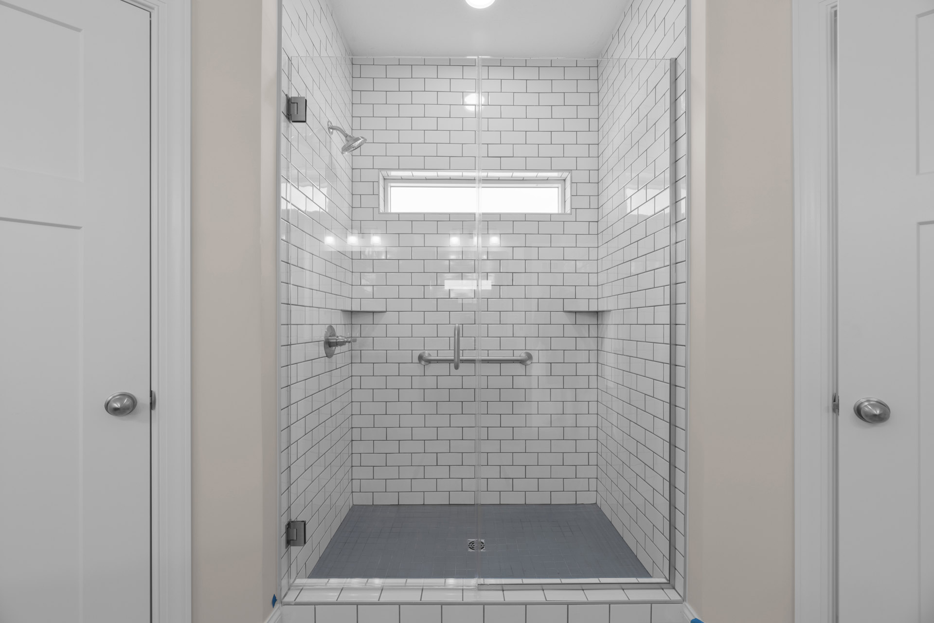 Frameless glass shower enclosure with chrome door handle, white tile walls, and rectangular window allowing natural light.