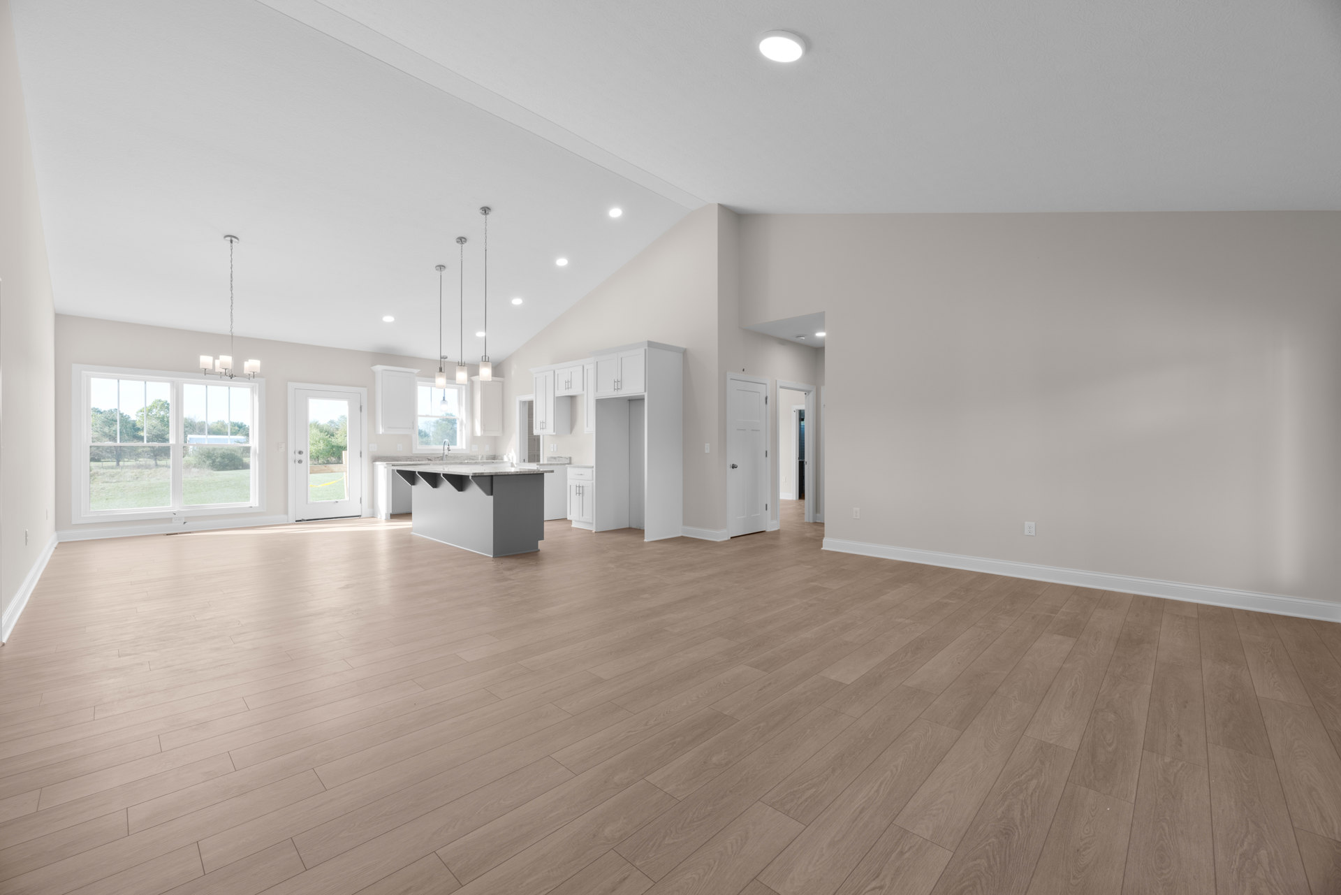 Spacious open floor plan featuring hardwood floors, white walls, kitchen island with sink, ceiling light fixture, white door and window overlooking green lawn and trees