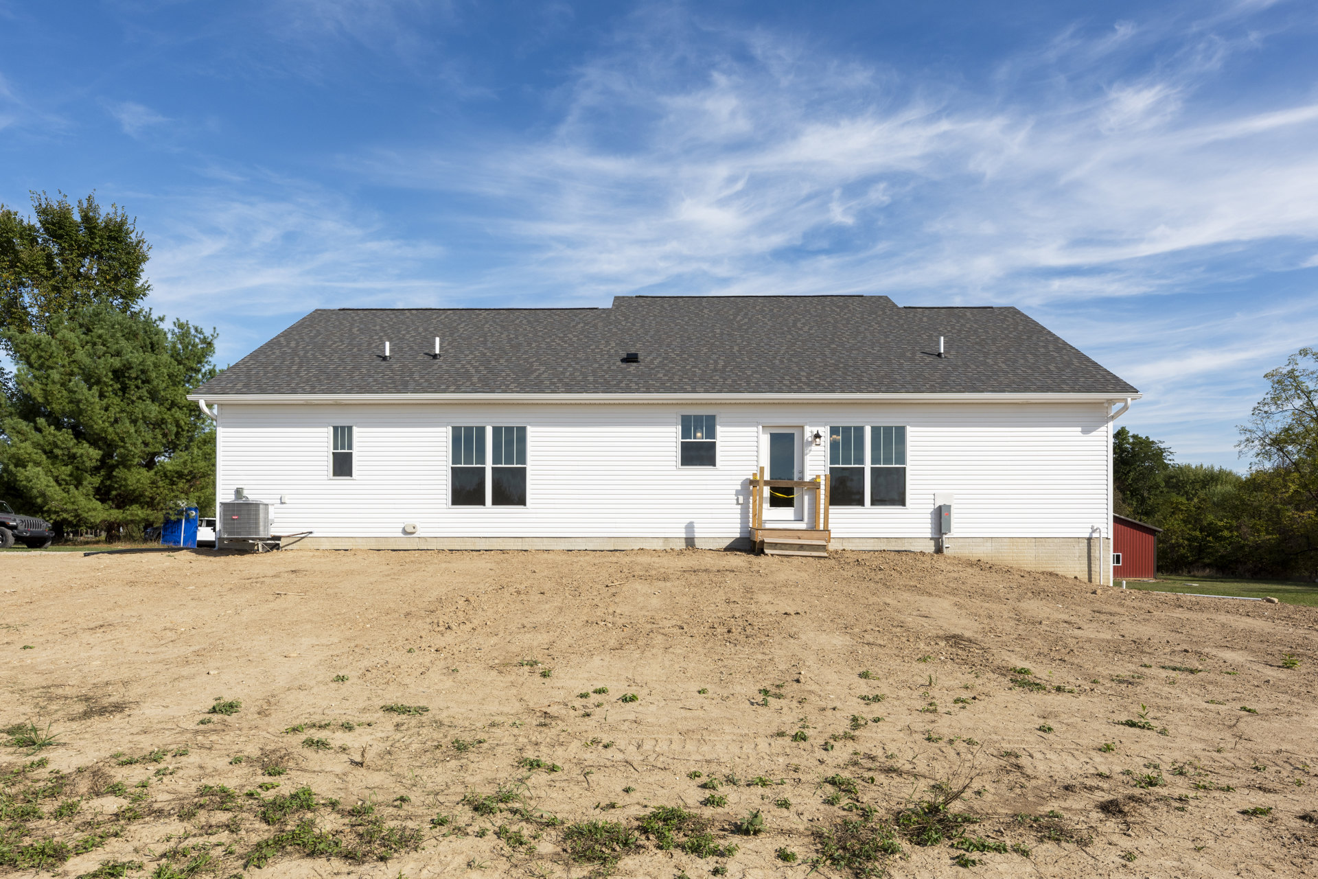 White house with covered porch and wooden deck, illuminated window, wooden steps leading to white door, dirt yard with scattered plants, parked car partially visible in foreground