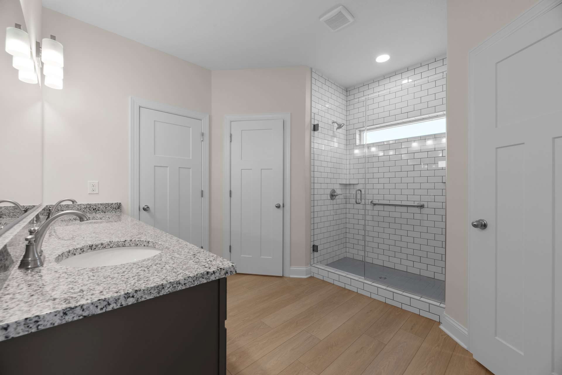 Bathroom with marble countertop, undermount sink and chrome faucet, glass-enclosed shower, white door with silver knob, modern light fixture, neutral cabinetry and tile flooring