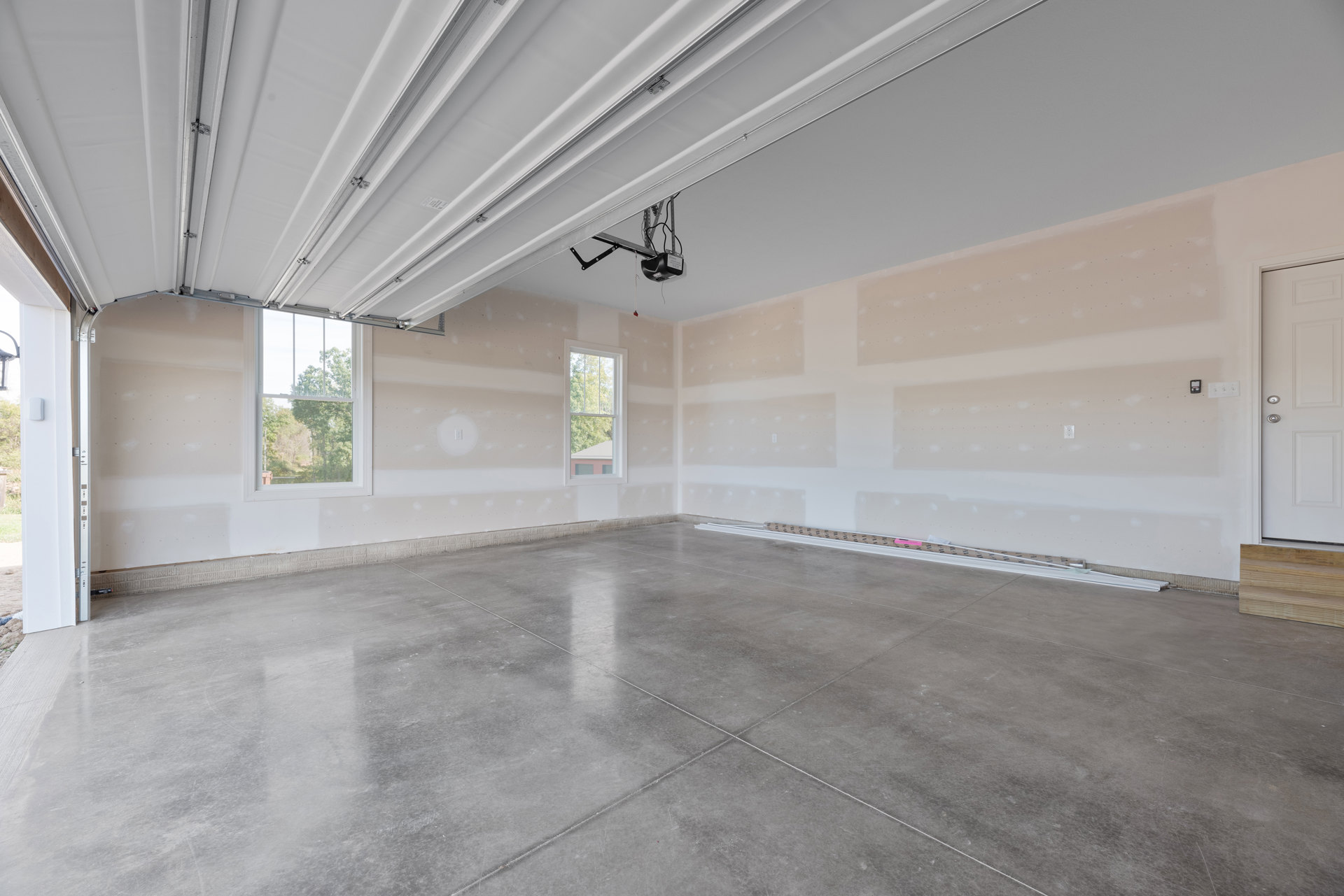 Empty room with smooth concrete floor, white plaster ceiling, white door, electrical outlet on wall, and window overlooking trees
