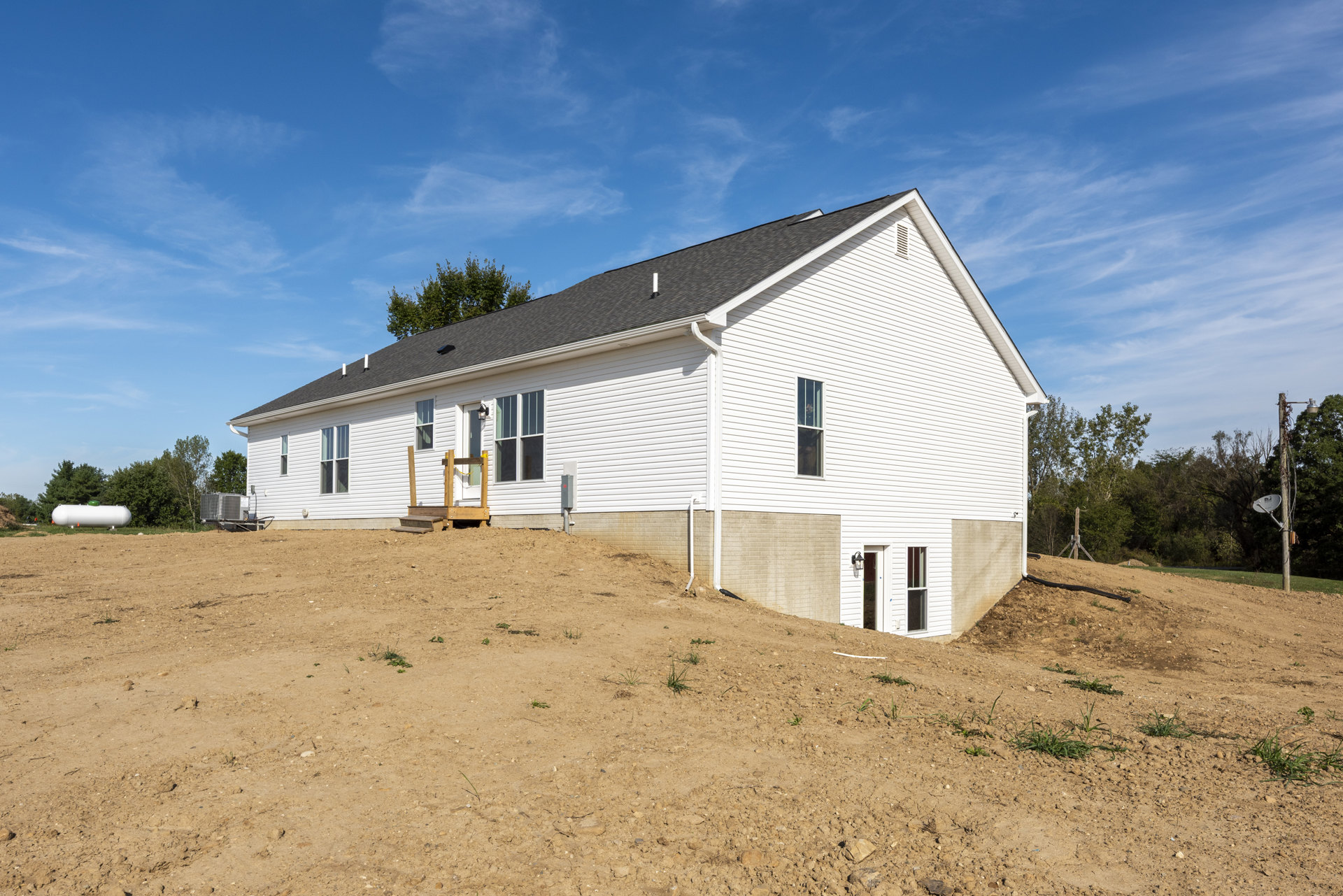 White house with black roof and wooden deck, set in front of a dirt hill with trees and cloudy sky in the background