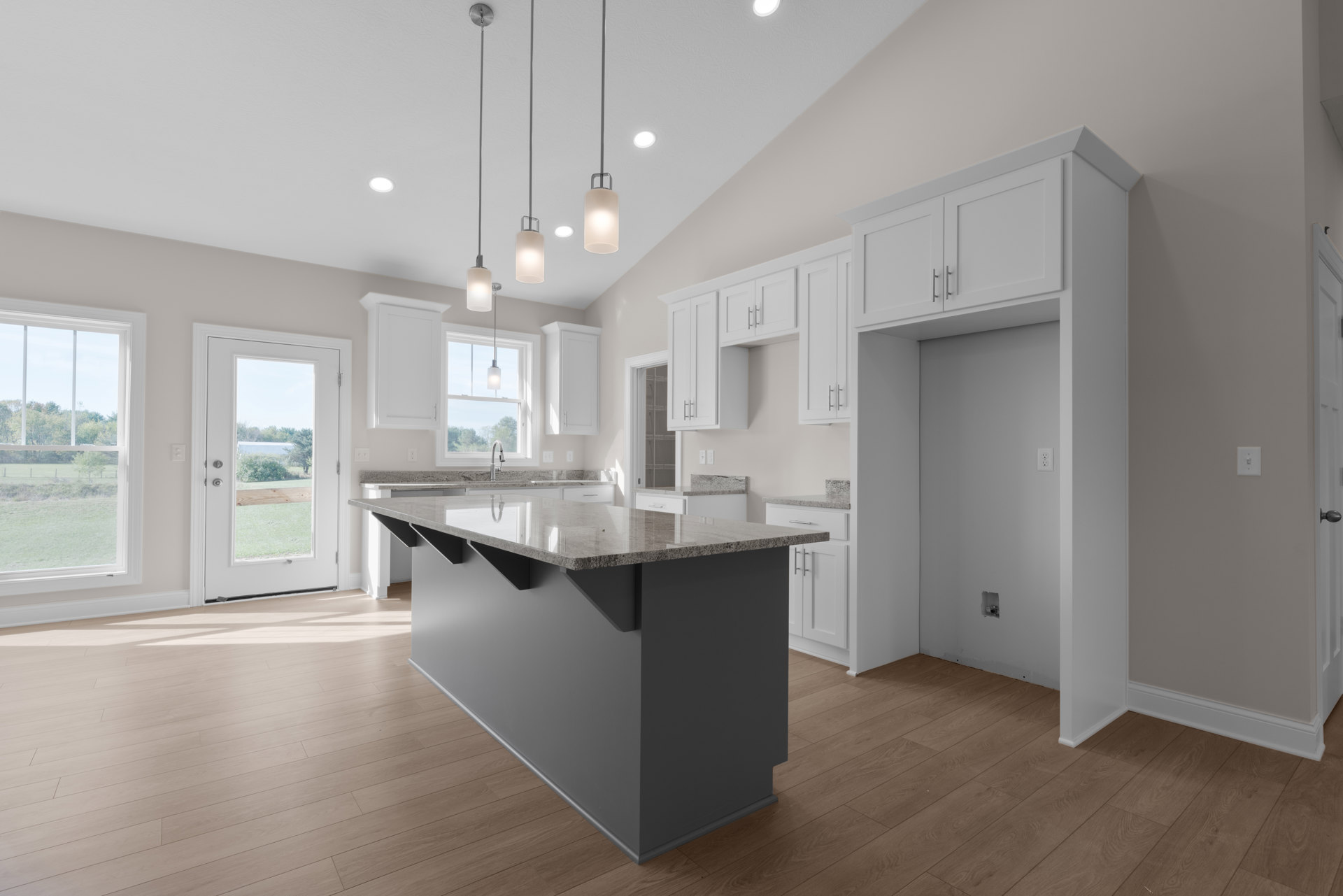 Spacious kitchen featuring a large marble island, white cabinetry, tile flooring, wall-mounted light fixture, window overlooking trees, and a door opening to the yard.