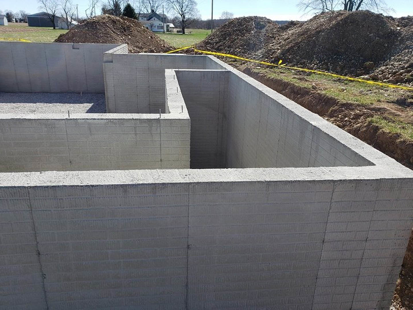 Concrete foundation with exposed hole, yellow caution tape on dirt pile, scattered rocks, blurred tree, distant building, and concrete wall marked with tape