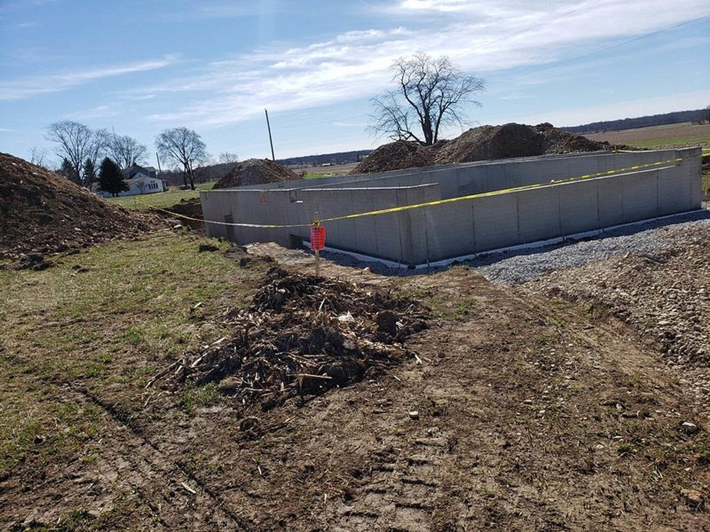 Concrete foundation wall marked with yellow caution tape, leafless tree nearby, pile of dirt and grass beside metal fence, overcast sky in background