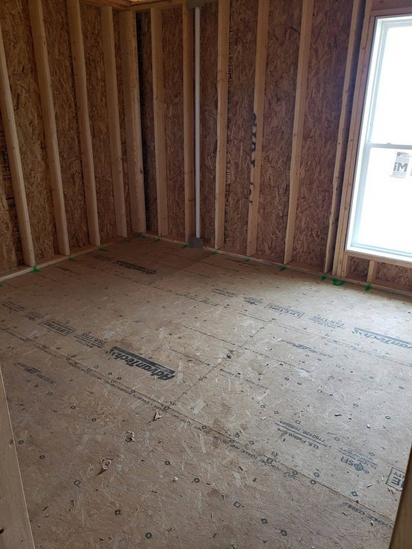 Room under construction with exposed wood framing, hardwood flooring, large window, white door featuring a sign, and a black mark visible on a grey surface.