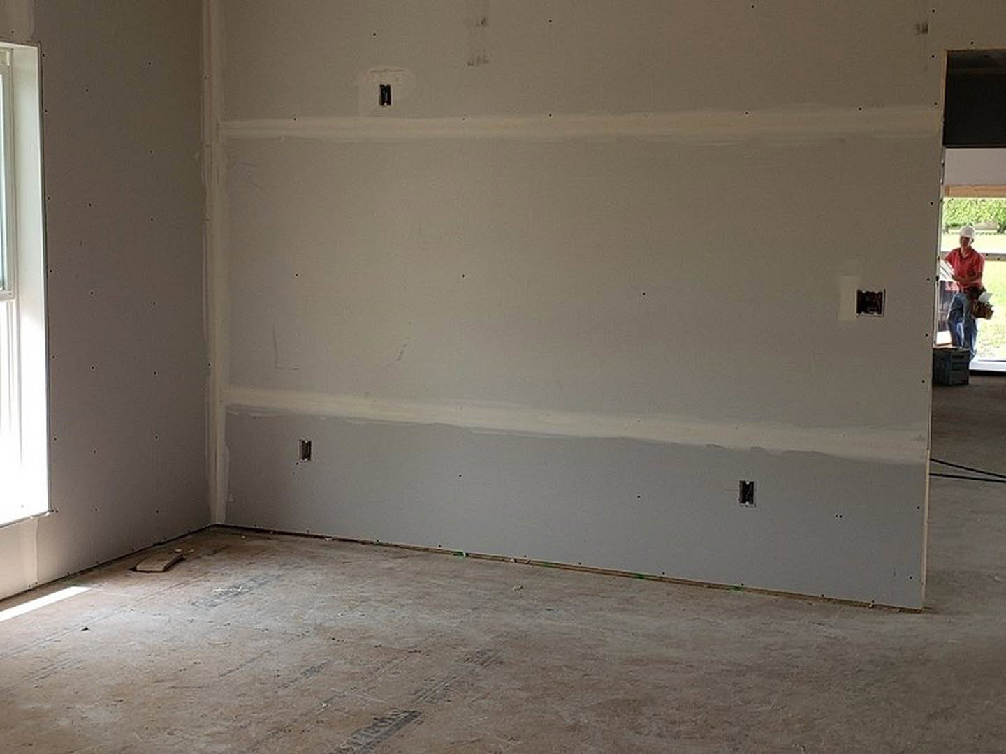 Sunlit room with white plaster walls, concrete flooring, window, door, and visible light switches and wall outlet