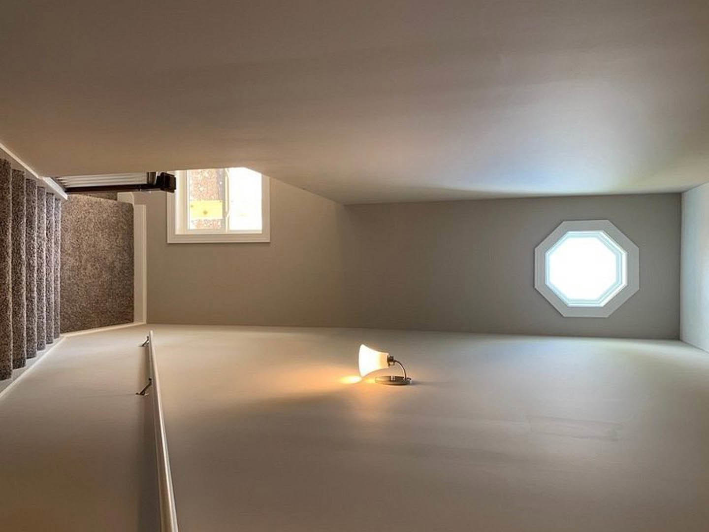 Track lighting fixture mounted on white plaster ceiling above a minimalist room with smooth walls and a large window.