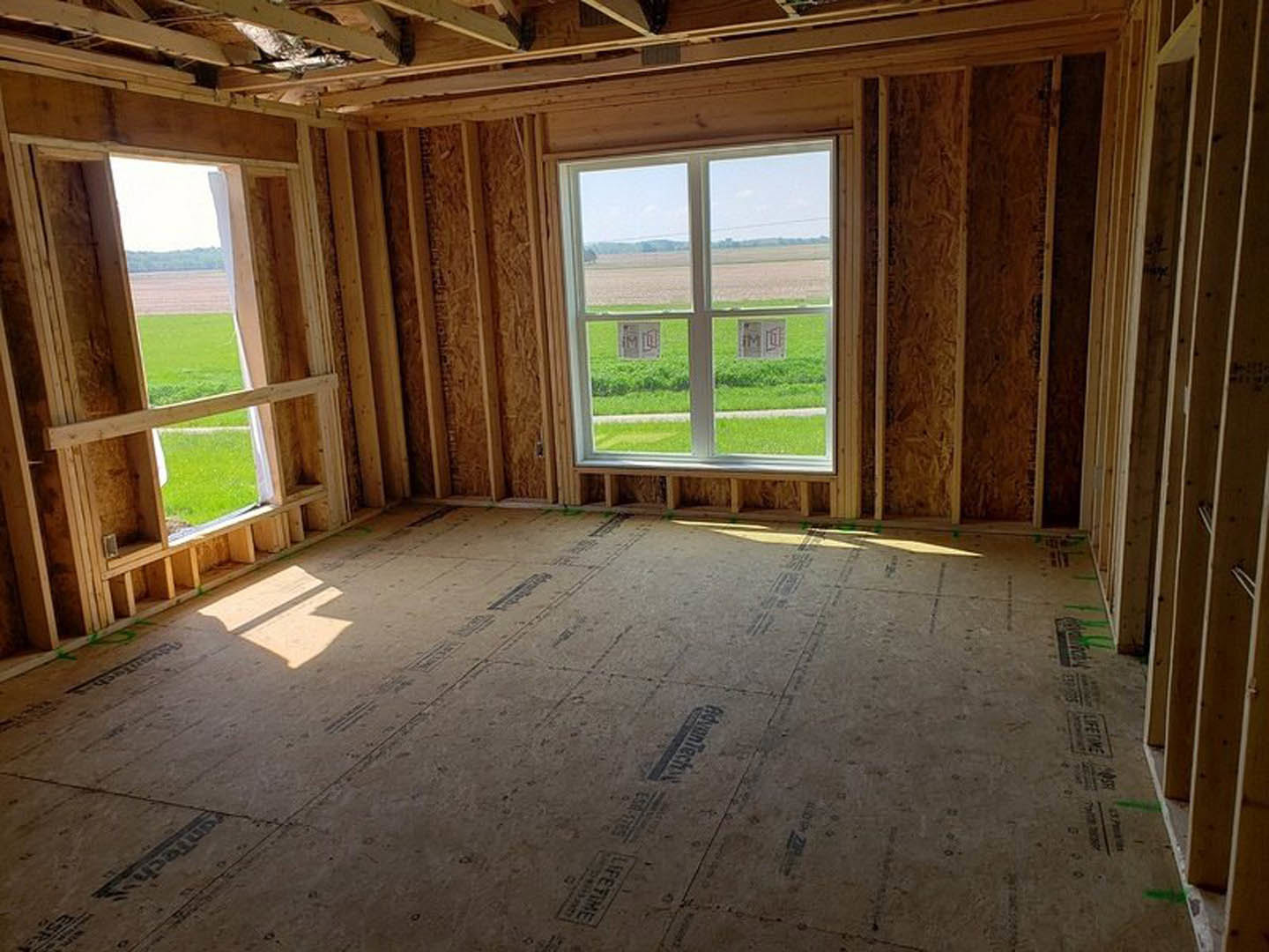 Sunlit room with wood plank flooring, large window overlooking grassy field, exposed ceiling beams, and daylight streaming across the floor.