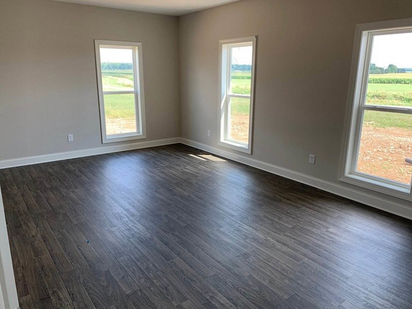 Sunlit room featuring large windows, natural hardwood flooring, white walls, and a view of grassy field outside.