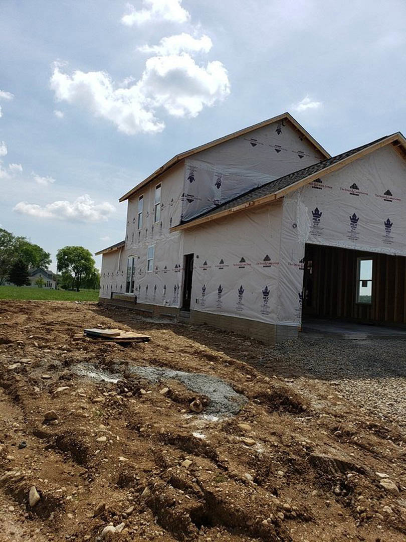 Two-story house under construction with plastic sheeting covering windows, exposed framing, and a dirt patch with a hole in the foreground; mature trees and cloudy sky in the