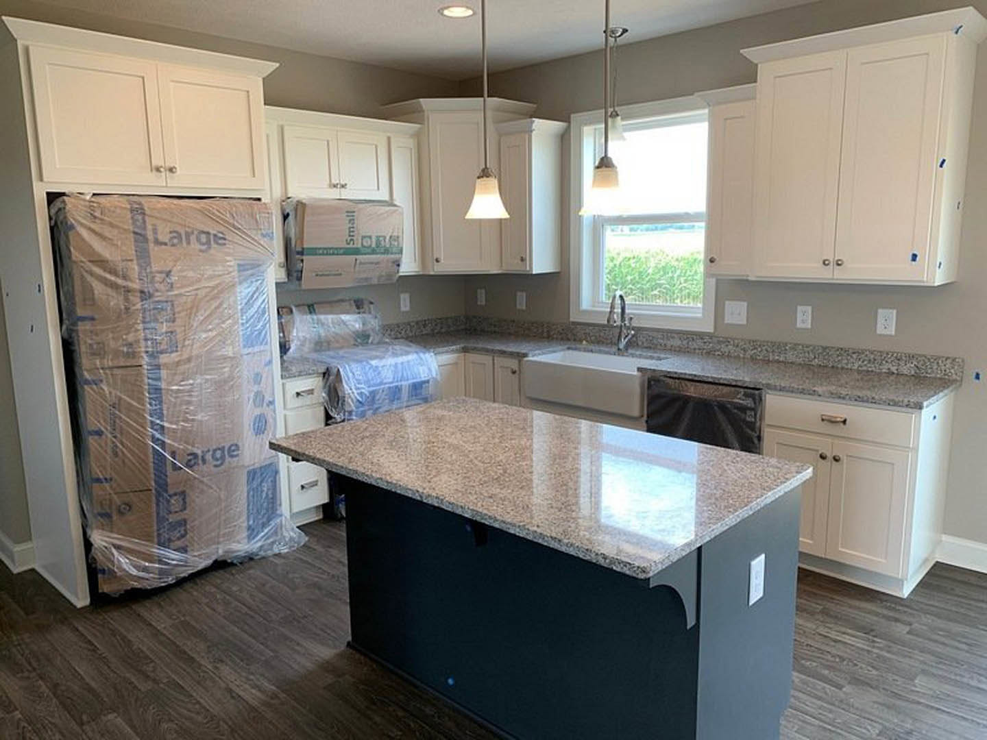 Kitchen with white cabinets, blue tape accents, and a large box wrapped in plastic on the countertop; light fixture visible near window.