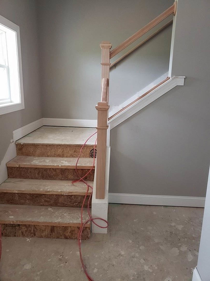 Staircase with wood steps and railings, white baseboard with red cable, large window letting in natural light, close-up of white pillar, plaster walls and hardwood flooring