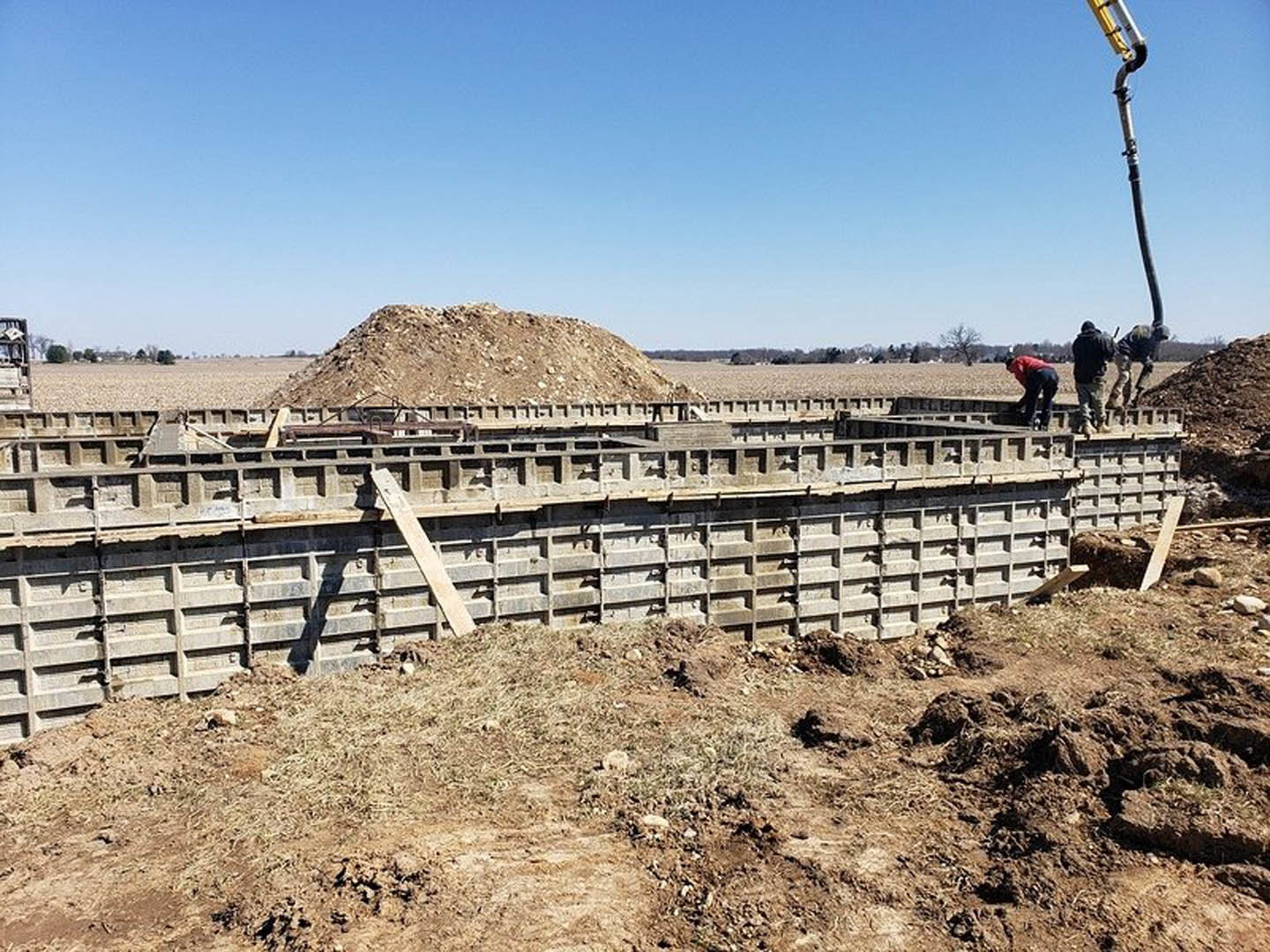 Construction site with exposed soil, unfinished foundation wall, man in black shirt and tan pants standing on concrete, blue sky overhead