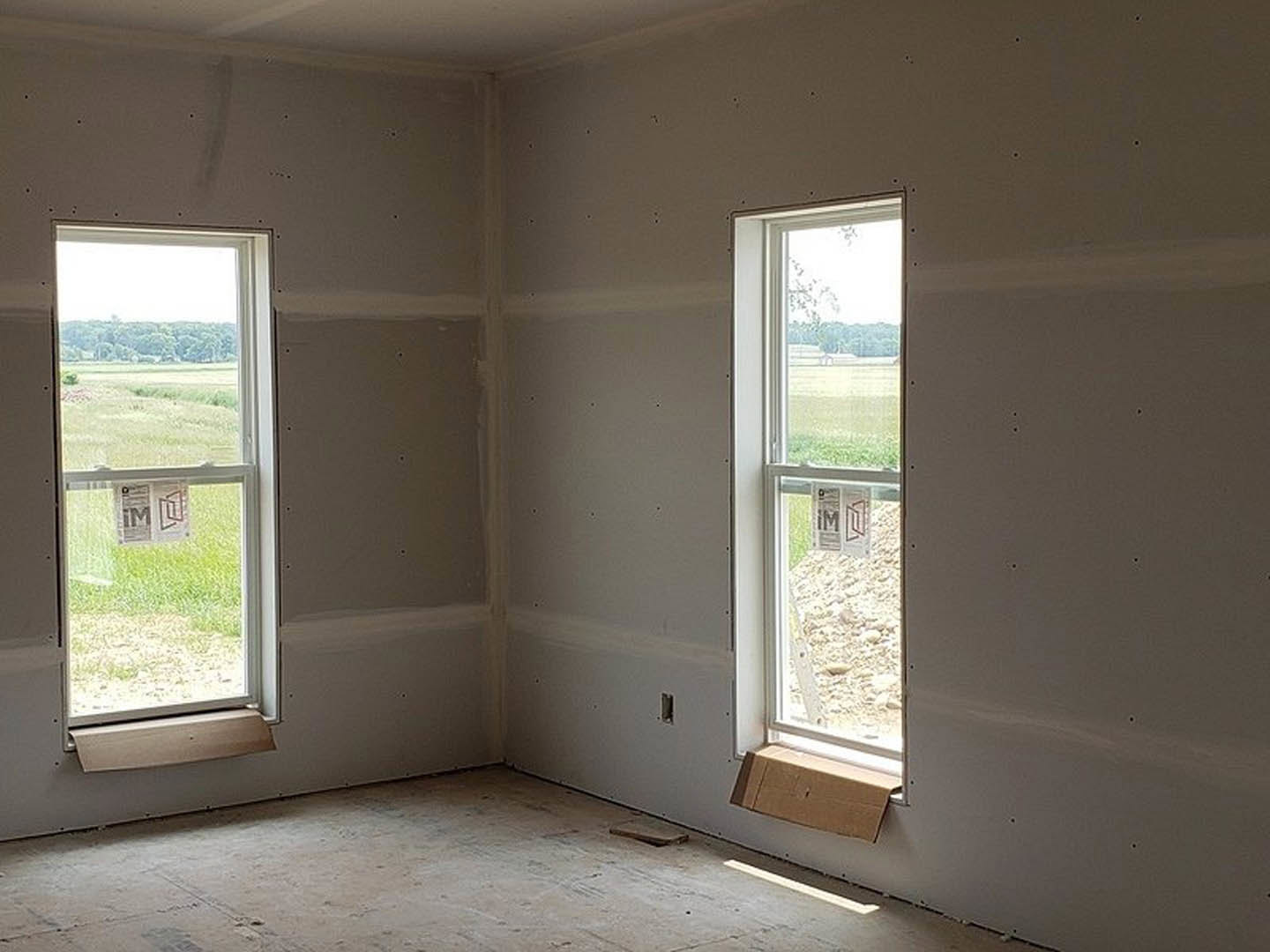 Bright room with two large windows, plaster walls, concrete floor, and a box placed near the wall; one window displays a newspaper, the other a sign.
