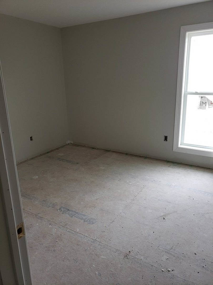 Bright room with white plaster walls, large window, light wood flooring, and white door frame with visible hardware holes