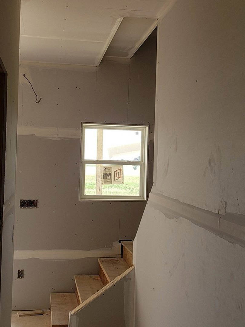 Unfinished wooden staircase with exposed framing, white plaster walls, cracked section near base, large window covered with newspaper, construction materials scattered on floor
