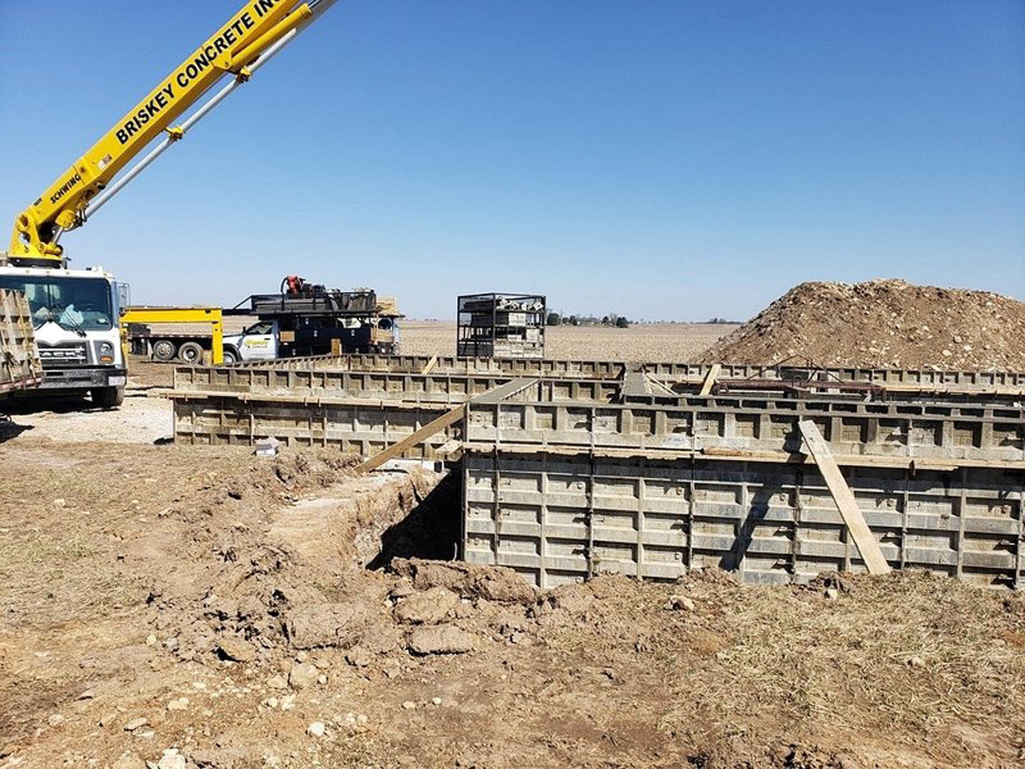 Framed custom home under construction with exposed wooden beams, crane lifting materials, soil-covered ground, and construction vehicles parked nearby