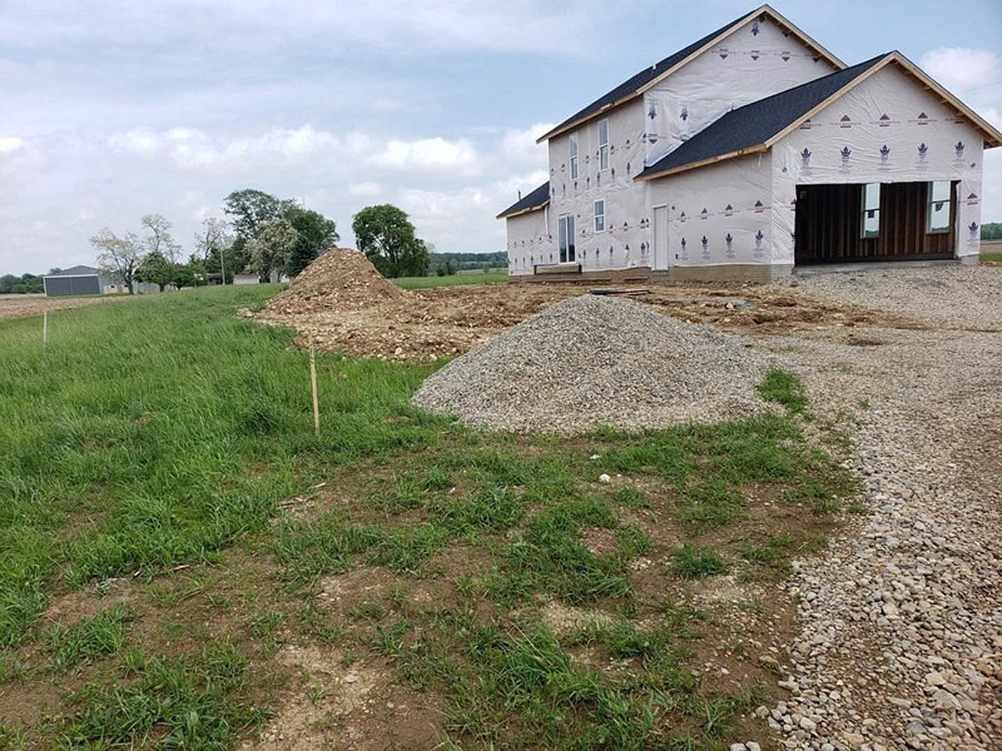 White farmhouse with black roof, square windows, attached garage, piles of gravel and dirt on grassy lot, cloudy sky overhead