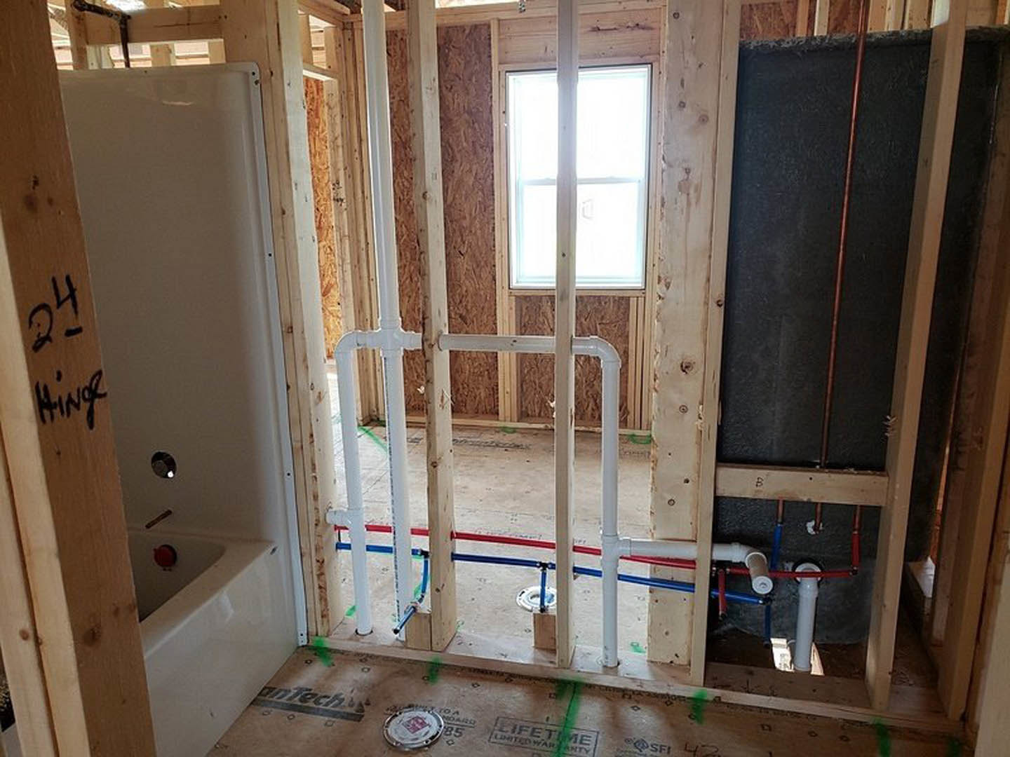 Bathroom under construction featuring exposed wood framing, white plumbing pipes, unfinished walls, bathtub, shower area, and window letting in natural light