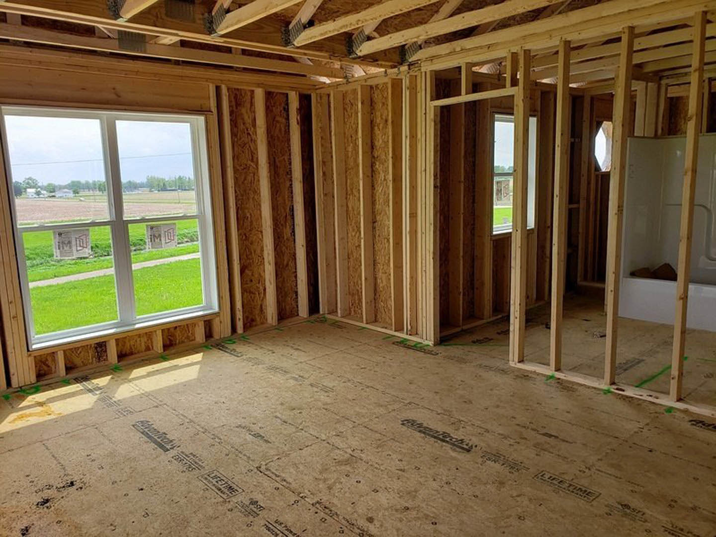 Wood-framed room under construction with exposed beams, unfinished floor, and large window overlooking grassy field