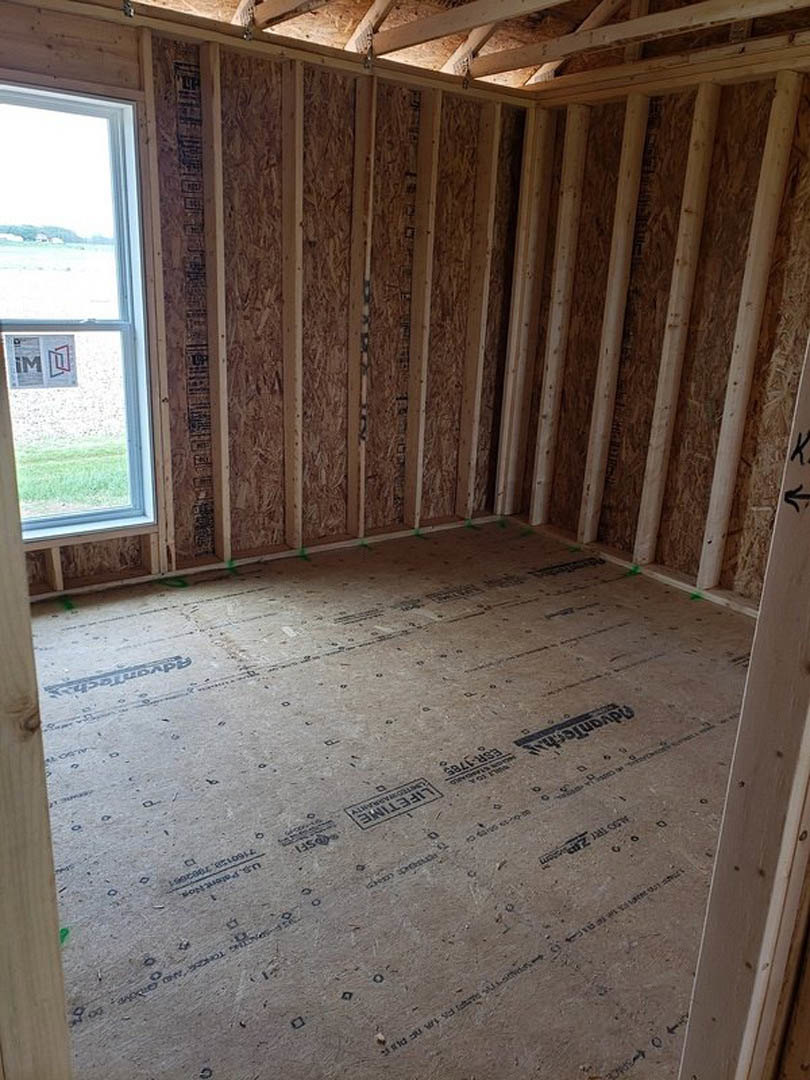 Unfinished room with exposed wood framing, plywood floor, roof trusses, and a window displaying a sign
