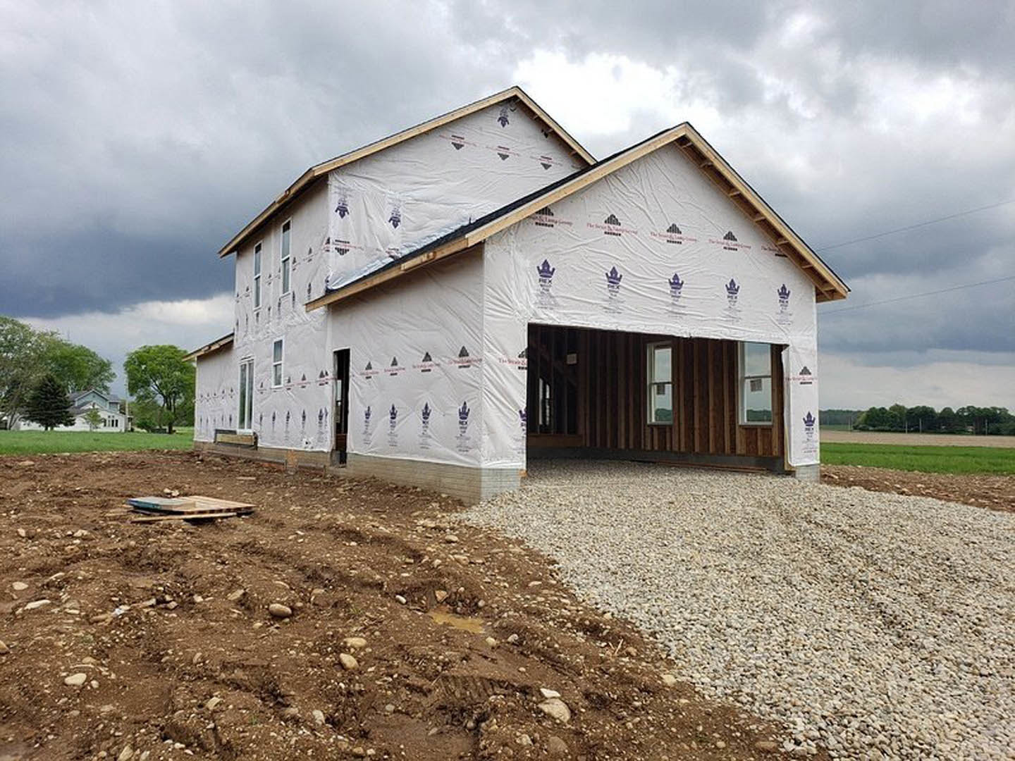 Partially built house with exposed framing, plastic sheeting covering windows, gravel and dirt piled along unfinished walkway, cloudy sky overhead, trees in background
