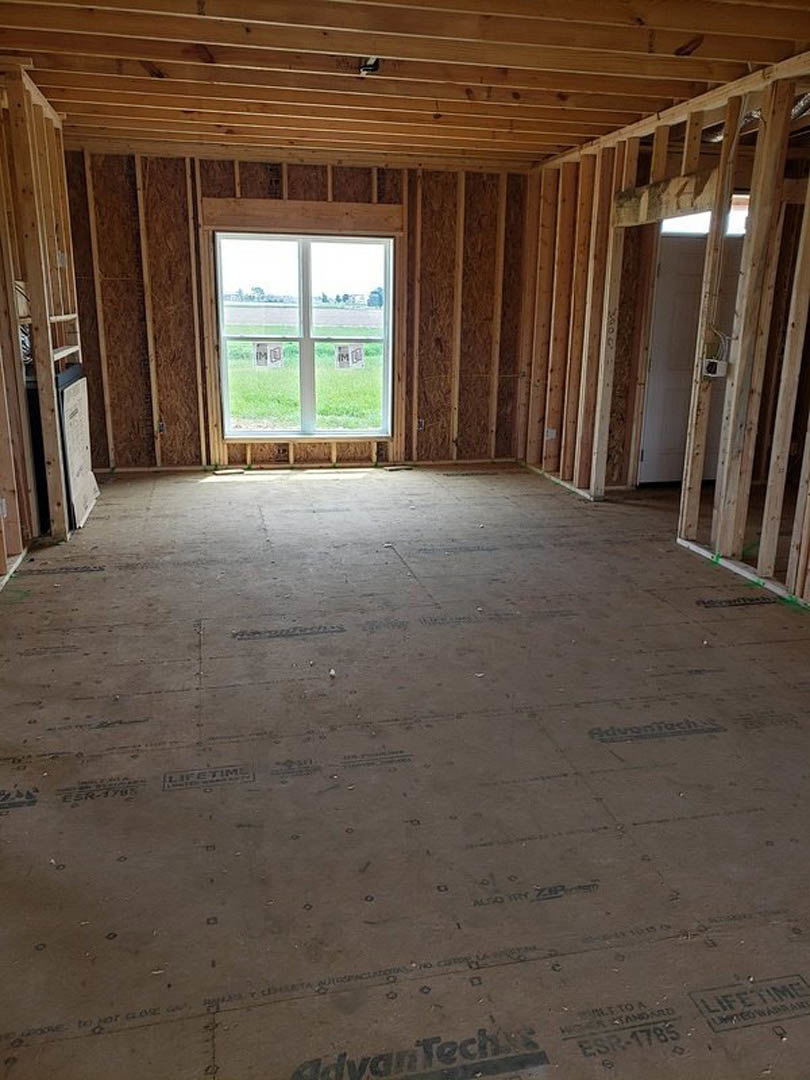 Room under construction featuring exposed wood framing, ceiling beams, and a window with posted signs; unfinished flooring and visible building insulation.