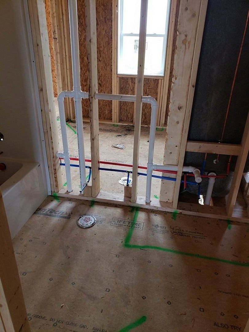 Bathroom under construction with exposed white pipes, unfinished plaster walls, wooden ceiling beam, window, and green line marked on concrete floor