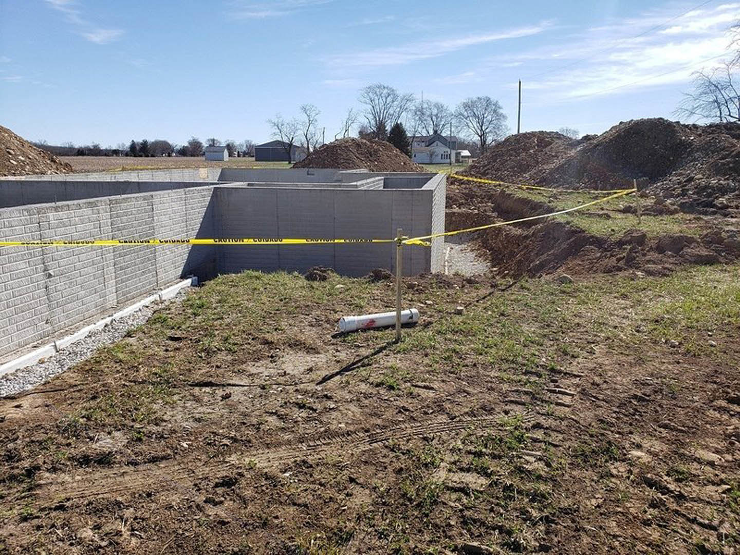 Dirt construction site with tire tracks, brick wall close-up, white pipe with red stains, pile of brown grass, wire fencing, trees under blue sky with clouds