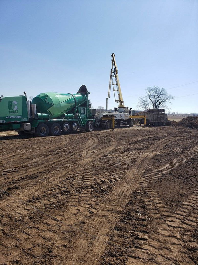 Construction site with green cement mixer truck, yellow crane, leafless tree, dirt ground with tractor tracks, and unfinished custom home exterior