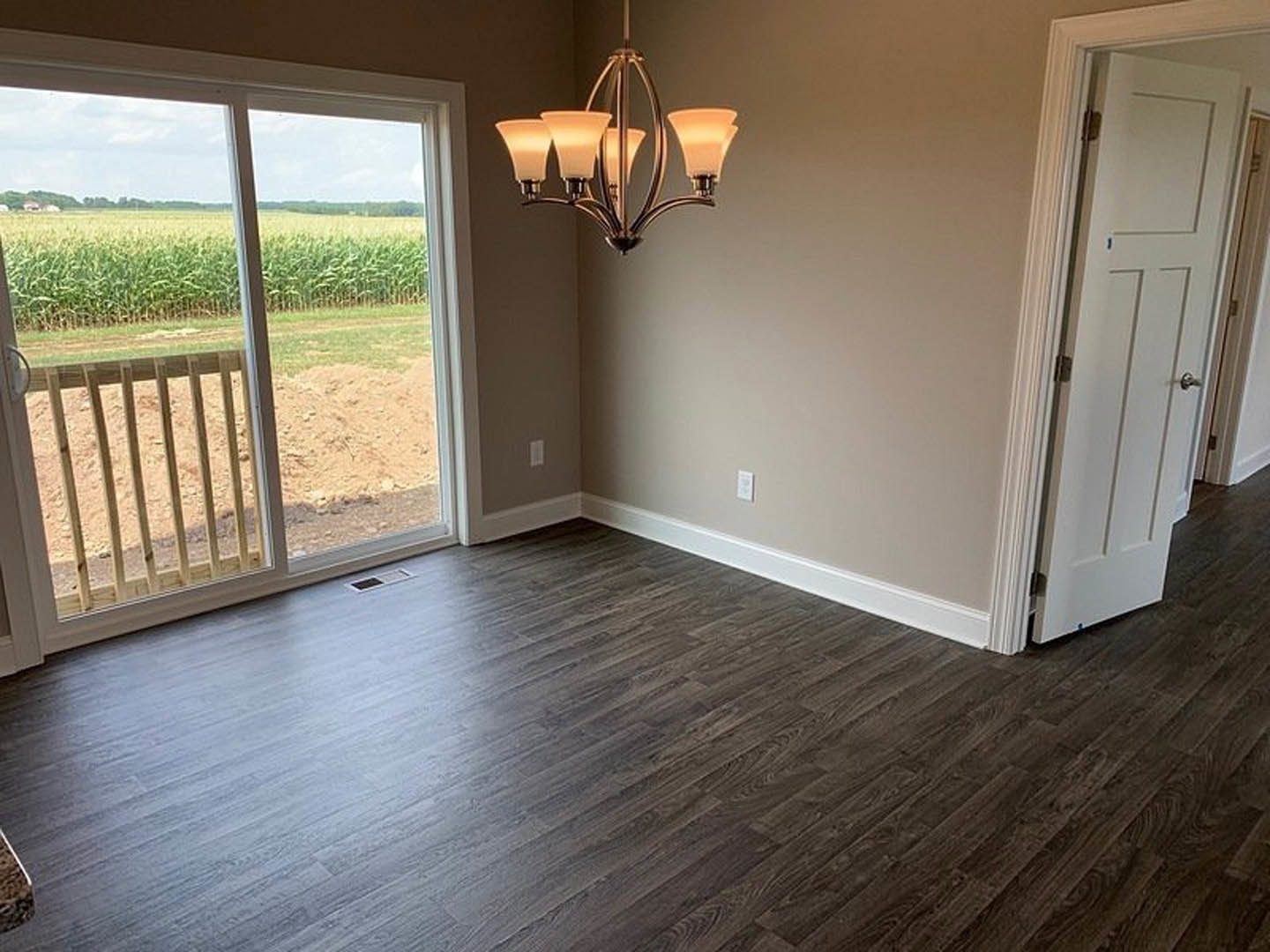 Dark wood flooring in a room with a white door featuring silver knobs, glass door, window overlooking a corn field, close-up chandelier, and wooden railing visible outside the