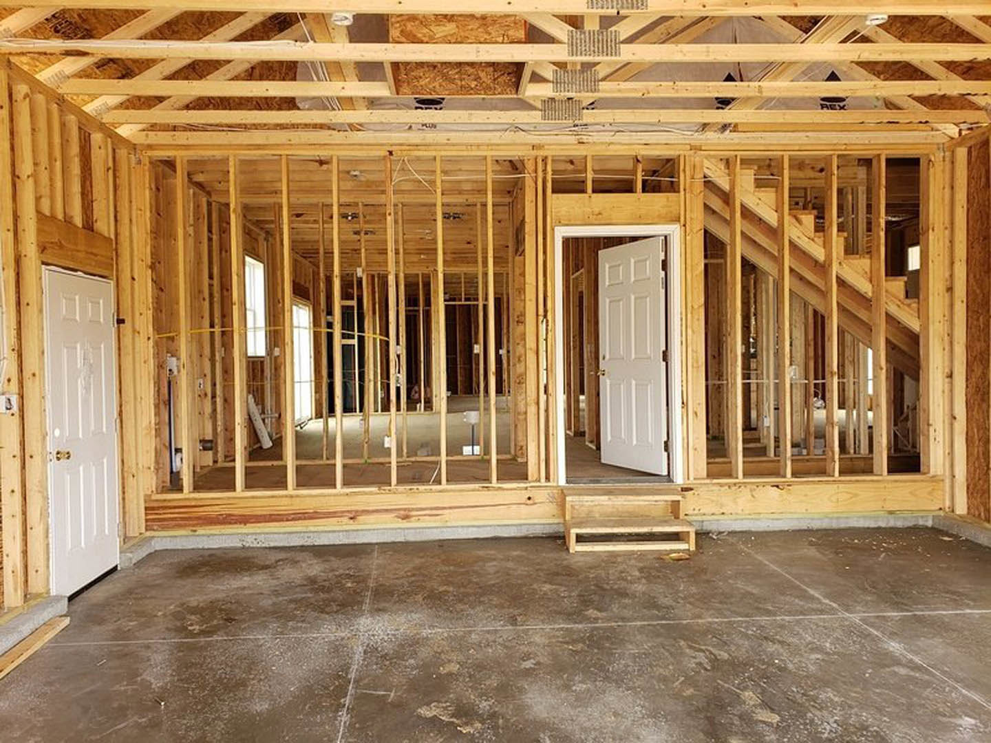 Unfinished interior with exposed wooden framing, white door featuring gold handles, concrete floor, and partially built staircase