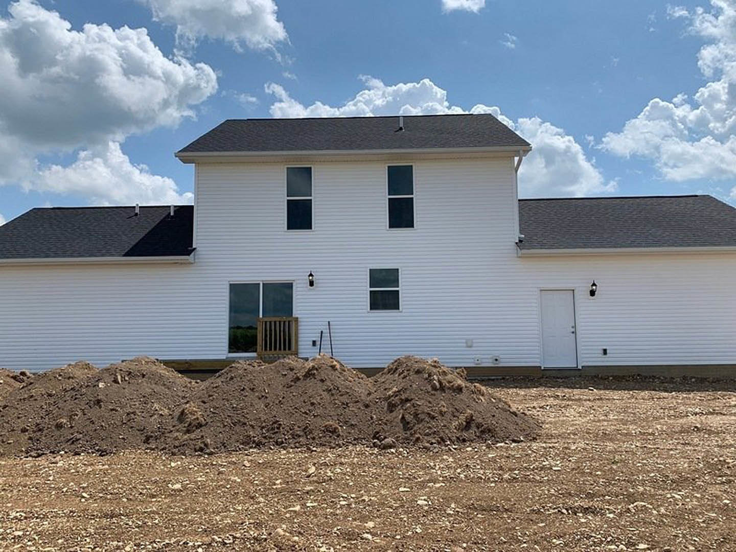 White house with horizontal siding, wooden porch railing, white front door, and a pile of dirt in the yard, partially enclosed by a wooden fence.