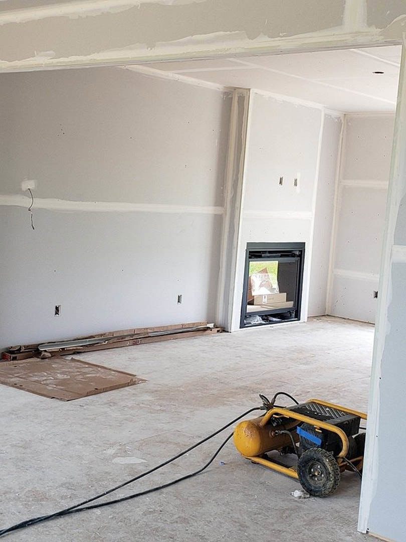 Yellow and black air compressor on unfinished floor near glass door, white plaster walls, exposed cable, cardboard with white spots, and tire in remodeled room.