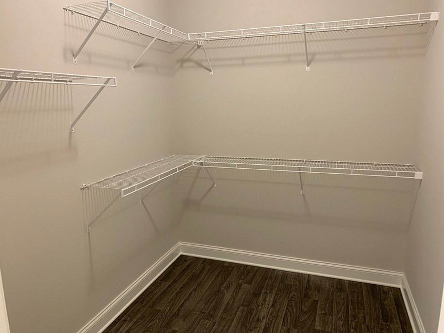 White built-in shelves and wire racks mounted on plaster walls, dark wood flooring with white baseboards, white clothes rack with railing, white metal grate on a shelf.