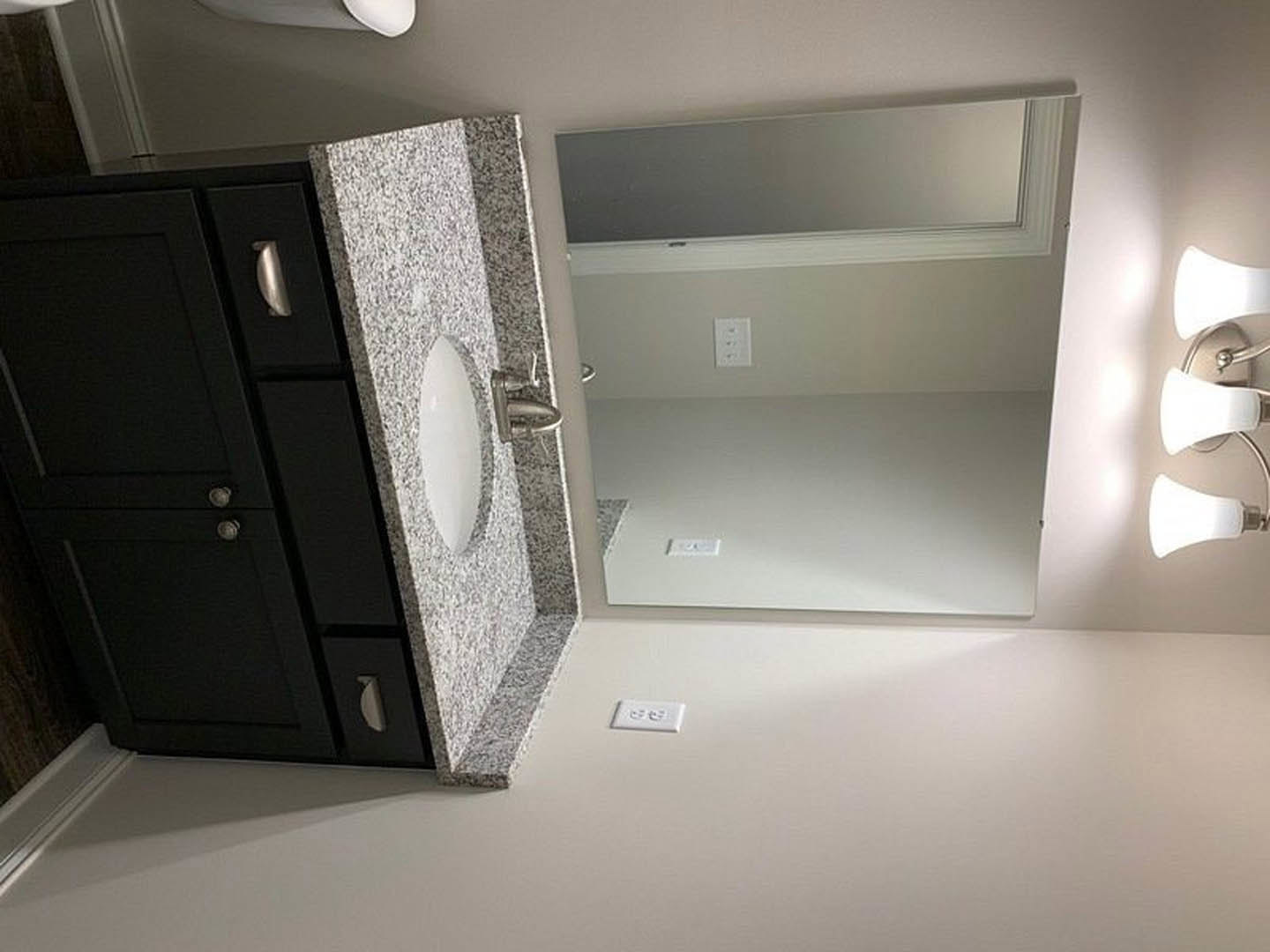 Modern bathroom featuring a rectangular mirror above a white sink, black vanity cabinet with silver handles, and white wall outlet with three switches on a light-colored wall