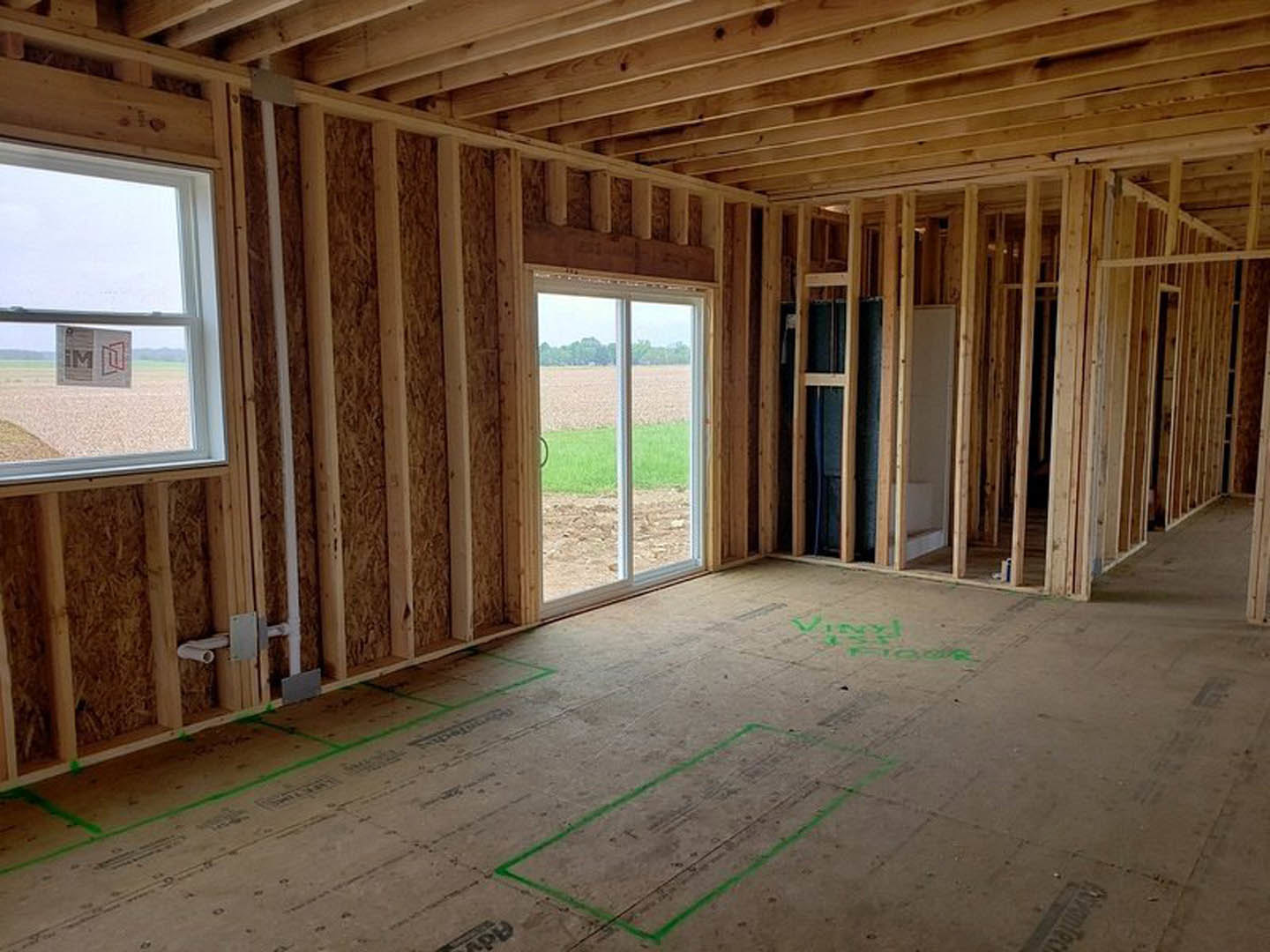 Living room with exposed wood ceiling beams, large windows overlooking a grassy field, light-colored walls, and hardwood flooring with green tape markings.