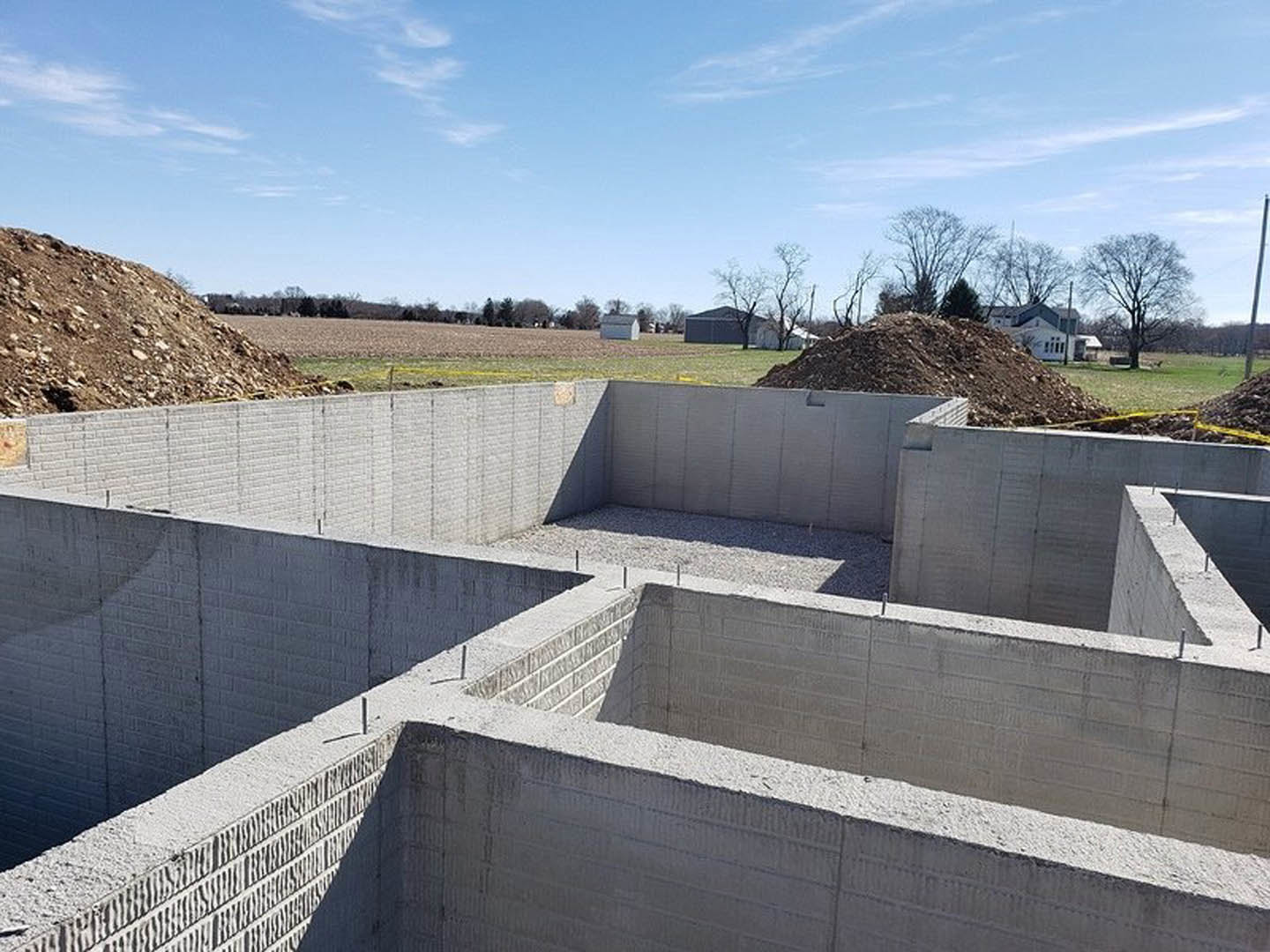 Concrete foundation with square openings surrounded by dirt piles, leafless tree, distant house, and partly cloudy blue sky