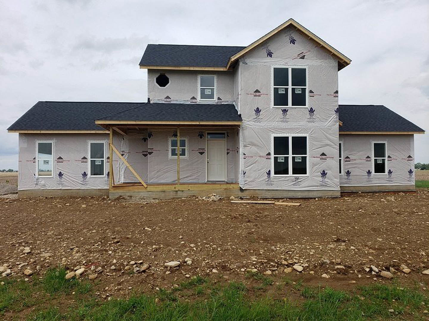 Partially built house with plastic sheeting covering exterior, white framed windows, white entry door with doorknob, dirt and grass in foreground, cloudy sky above